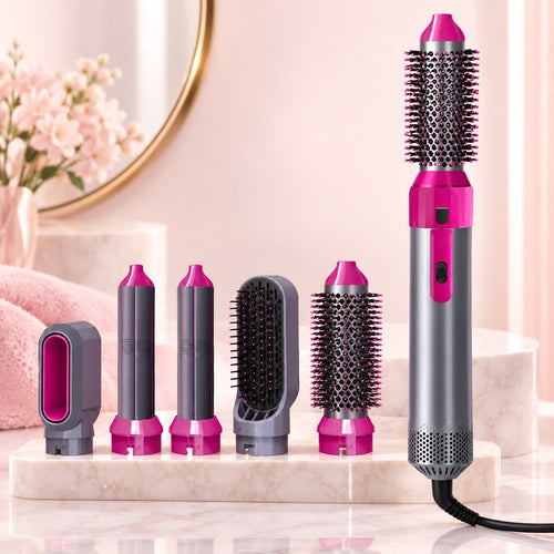A hair styling tool set with a tall round brush attachment standing upright on the right and four smaller attachment heads lined up on a marble pedestal to the left. The tools are primarily gray and bright pink/magenta, photographed in a softly styled interior with a mirror, flowers, and pink decor in the background.