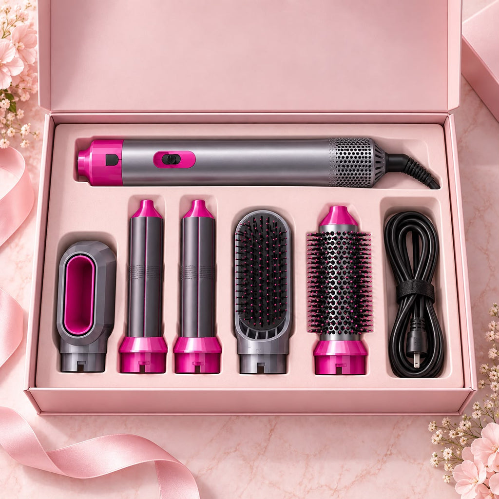 A hair styling tool set arranged in an open pink presentation box, including a main styler, three barrel attachments, a flat brush attachment, a round brush attachment, and a black charging cable. The tools are primarily silver and dark gray with bright pink accents, set against a soft pink marble background with decorative flowers and ribbon.